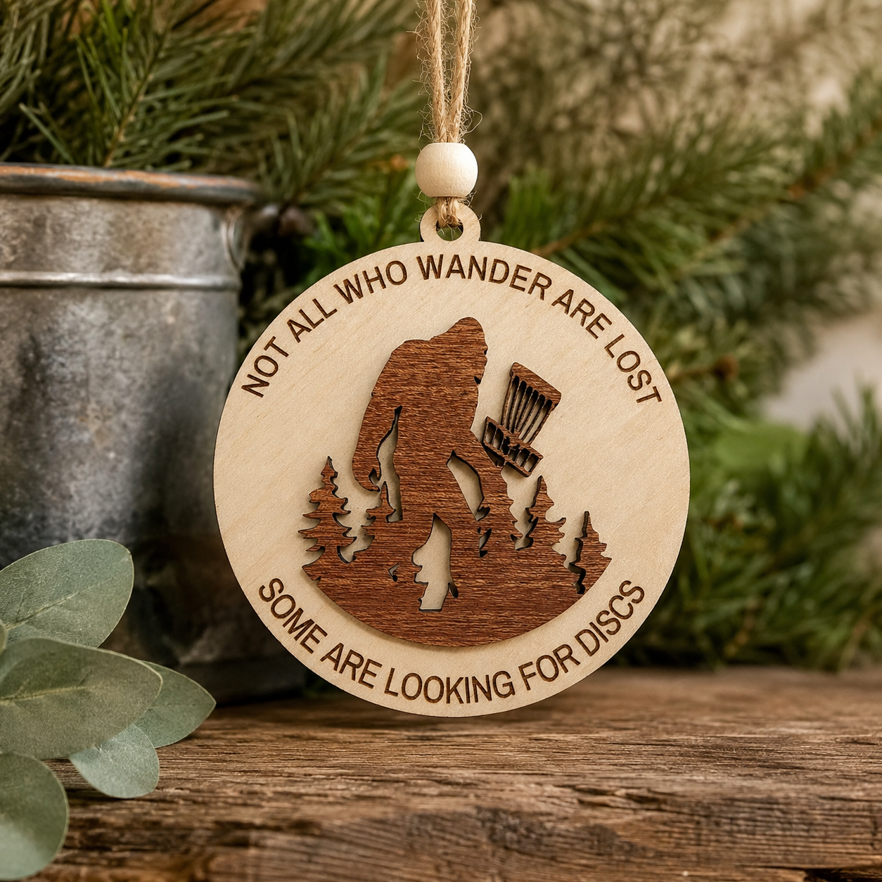 Handmade layered wood Bigfoot disc golf ornament hanging in a rustic holiday setting with jute twine and wooden bead.