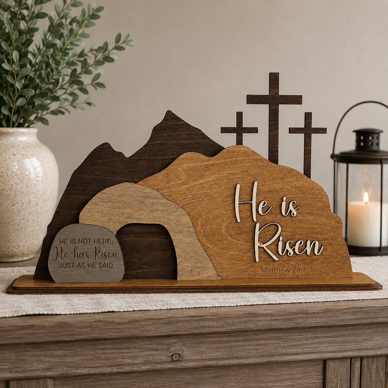 He Is Risen wooden resurrection scene displayed on a rustic farmhouse table with greenery and candle, handcrafted Christian Easter shelf decor featuring the empty tomb and three crosses.